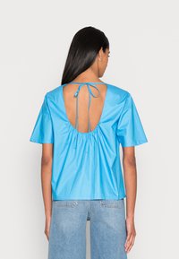 Top de manga curta, azul claro, com uma grande abertura nas costas e um detalhe de cordão. Feito de um tecido suave com textura franzida à volta do decote.