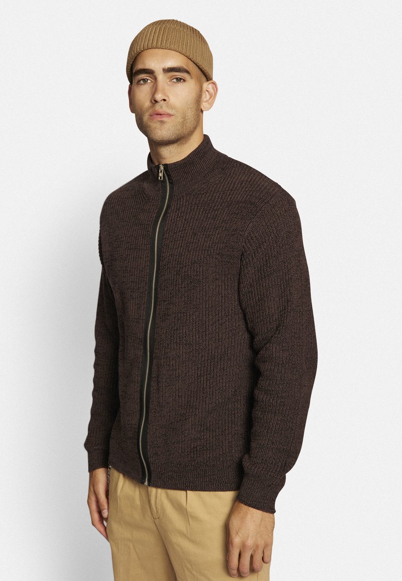 Brun strikket lynlås sweater med høj krave og ribbet tekstur. Har en guldlås og tætsiddende ærmer, parret med beige bukser.