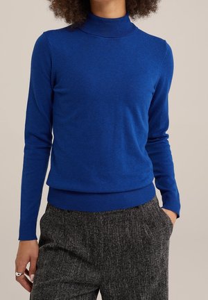 Personne portant un pull à col roulé bleu roi ajusté à manches longues et un pantalon gris texturé, avec une main dans la poche et une bague en argent.