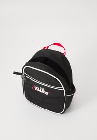 Rucsac din material negru, cu detalii albe, accent cu fermoar roșu și logo brodat alb "nike". Dispune de un mâner superior și două închideri cu fermoar.