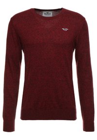 Punainen neulepaita klassisella pyöreällä pääntiellä, pitkät hihat ja pieni valkoinen logo rinnassa. Pehmeä rakenne ja hieman istuvampi muotoilu.