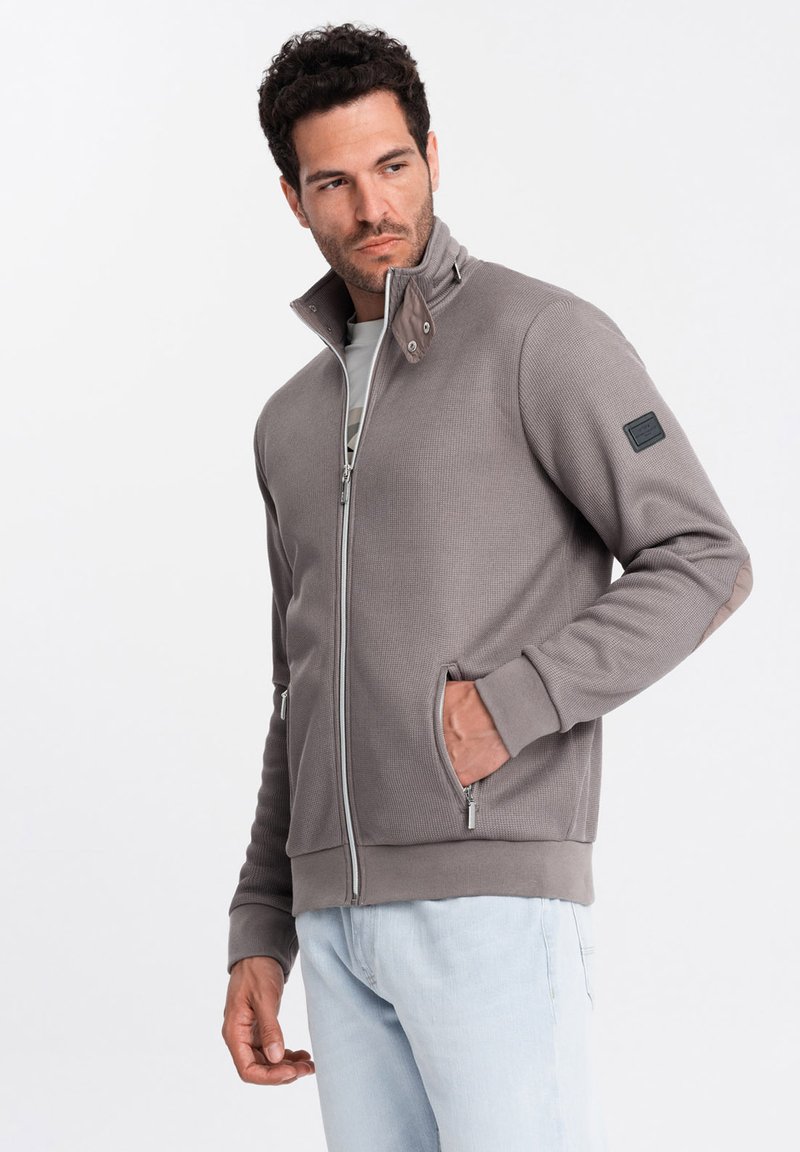 Giacca grigia con zip e un alto colletto, texture a coste e due tasche frontali. Presenta un tessuto a contrasto sul colletto e sulle spalle.
