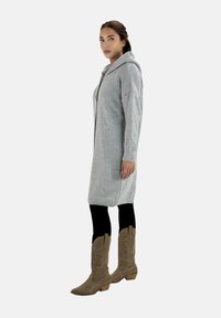 Helle graue Strickjacke mit Kapuze, locker geschnitten und mit gerippter Struktur, getragen über schwarzen Leggings und hellbraunen Cowboystiefeln.