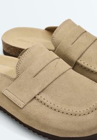 Sabots beige en daim à enfiler avec une bride cousue et une semelle en liège sur fond blanc.