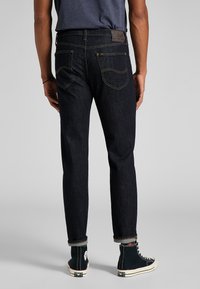 Jeans de mezclilla oscura de pierna recta, con costuras amarillas, un parche de cuero en la parte trasera y dobladillos enrollados sobre zapatillas negras de caña alta.
