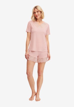 Vrouw staat blootsvoets in een zachtroze T-shirt met korte mouwen en bijpassende korte broek met kanten afwerking aan de halslijn en zoom van de shorts.