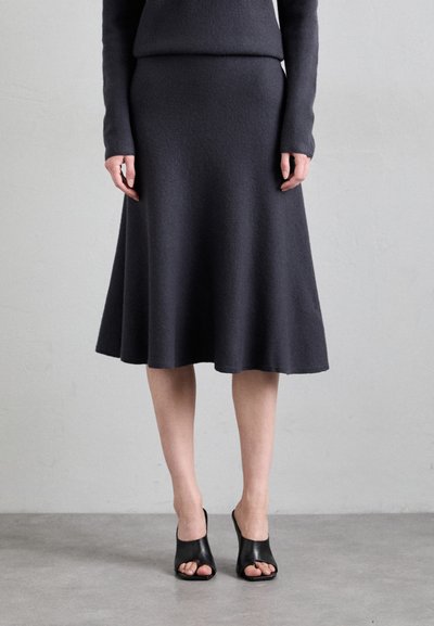 Jupe trapèze grise foncée, longueur genou, en tissu doux, associée à des talons ouverts noirs. Lignes simples et épurées avec une finition texturée.