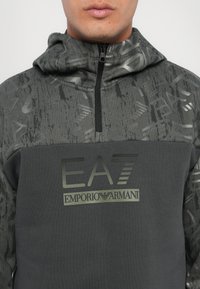 Grå zip-up hoodie med et tekstureret mønster, der har et mørkt brystpanel og EA7-logo. Materialet virker blødt og atletisk.