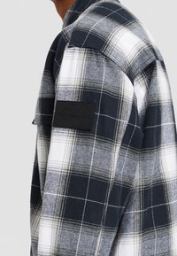 Camicia di flanella a quadri bianco e nero con una texture morbida, caratterizzata da una toppa con logo nero lucido sulla parte superiore del braccio e un colletto classico.