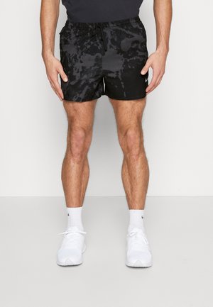 Mann trägt schwarze, gemusterte Sportshorts, weiße Nike-Socken und weiße Turnschuhe, steht vor einem schlichten hellen Hintergrund.