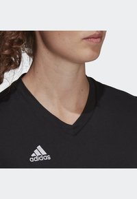 Tricou negru Adidas cu un design de guler în V. Prezintă un logo alb brodat pe partea stângă a pieptului. Material moale cu o textură fină.