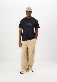 Black t-shirt with Quiksilver logo, beige wide trousers, and light gray cap. Shoes are light beige with darker accents, designed for comfort.