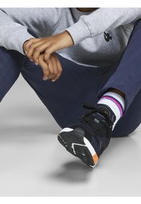 Persona con sudadera gris y pantalones azul marino sentada con las piernas cruzadas, usa zapatillas negras con acentos en blanco y naranja y calcetines blancos con rayas rosas y azules.