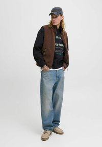 Junger Mann in einem braunen Fleece-Weste, schwarzem Sweatshirt, lockeren blauen Jeans, beigen Schuhen und einer dunklen Baseballmütze, steht mit Händen in den Taschen.