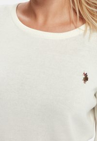 Pull crème clair avec un col côtelé, orné d'un petit logo brodé sur la poitrine. Le tissu semble doux et texturé.