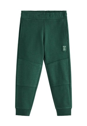 Ungt barn med blondt hår iført en beige kasket, brun- og hvidstribet langærmet skjorte, grønne bukser og hvide sneakers, står og smiler.