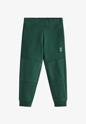 Junges Kind mit blondem Haar, trägt eine beige Mütze, braun-weiß gestreiftes Langarmshirt, grüne Hose und weiße Sneakers, steht und lächelt.