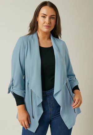 Cardigan drappe di un blu chiaro realizzato in tessuto testurizzato, con maniche a tre quarti e dettagli con fiocchi, e tasche frontali, indossato sopra una maglia nera.