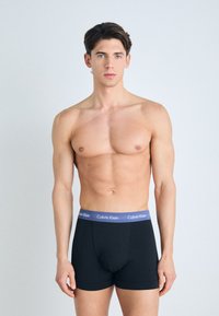 Fekete pamut boxer alsónadrág lila derékkal, melyen a "Calvin Klein" logó szerepel. A modell egy egyszínű háttér előtt áll.