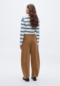 Gestreepte top met lange mouwen in wit en marineblauw; brede bruine broek met zijnaden; zwarte platte schoenen; golvend haar dat langs de rug valt.