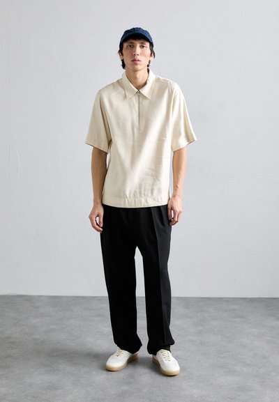 Chemise beige à col et manches courtes avec une demi-fermeture éclair, associée à un pantalon noir. Des sneakers blanches avec une semelle beige complètent la tenue.