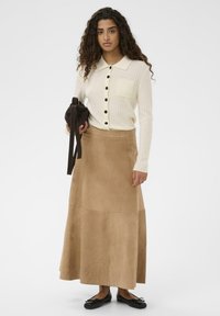 Gai krēmkrāsas ribota džemperis ar melnām pogām un kabatu, apvienots ar gaiši brūnu zamša maxi svārku. Modelis tur tumši brūnu somu.