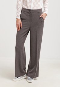 Pantalon gris à jambes larges en tissu lisse, doté d'une fermeture à bouton et de deux poches latérales, associé à des baskets argentées.