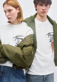 Δύο νέοι που φορούν λευκά μπλουζάκια με σχέδια, ο ένας με ένα χακί πράσινο μπουφάν περασμένο στα χέρια, ο άλλος με πράσινο πουλόβερ περασμένο στους ώμους.