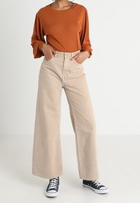 Rostfärgad långärmad topp som kombineras med högmidjade vida beige jeans och marinblå sneakers. Slät textur och enkel design med avslappnad passform.