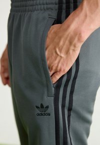 Pelēki sporta bikses ar melniem svītrainiem akcentiem, kas aprīkotas ar sānu kabatām un izšūtu melnu logotipu uz apakšējās kājas. Gluda, elastīga auduma struktūra.