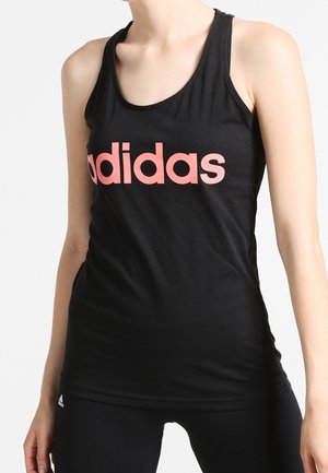 Vrouw die een zwarte mouwloze Adidas-top draagt met een roze logo en zwarte legging, staand tegen een effen lichte achtergrond.