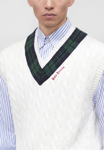Homme portant un gilet en maille torsadée blanc avec un liseré à carreaux verts et bleu marine, par-dessus une chemise rayée bleue et blanche à boutons.