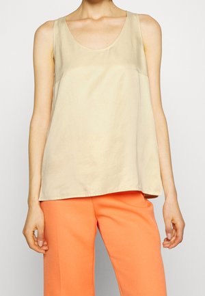 Femme portant un haut beige sans manches et un pantalon orange vif, montrée du cou jusqu'au milieu de la cuisse sur fond uni.