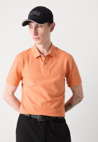 Narančasta polo majica s klasičnim ovratnikom i kratkim rukavima, uparena s crnim hlačama i crnim kapom s natpisom "BOSS" izvezenim bijelom bojom.