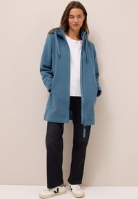 Veste à capuche en polaire bleue avec fermeture éclair à l'avant, deux poches latérales et cordon de serrage. Portée sur une chemise blanche et associée à un pantalon sombre et des baskets.