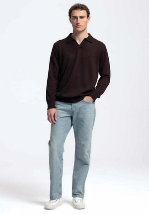 Jonge man staat met één hand in de zak van lichtblauwe jeans, draagt een donker lange mouw trui en witte sneakers op een witte achtergrond.