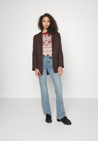 Blazer castanho sobre um suéter padrão vermelho e branco, combinado com jeans clarinhos de boca de sino e sapatos pretos, de pé contra um fundo neutro.