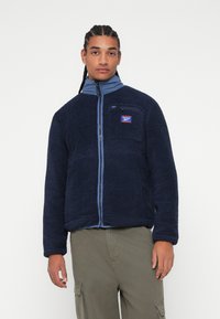 Fleece bunda v námořnické modři s vysokým límcem a zipovým uzávěrem, s náprsní kapsou a kontrastními světle modrými akcenty.
