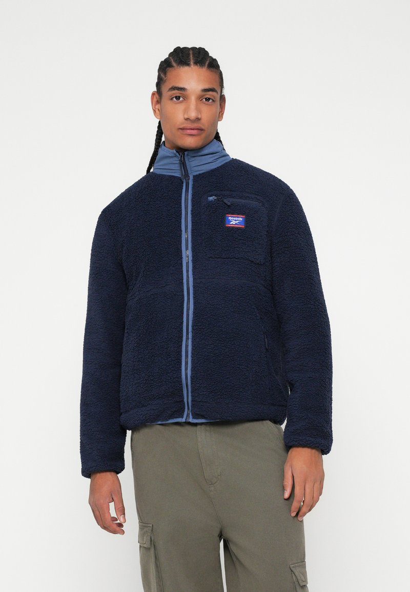 Fleece bunda v námořnické modři s vysokým límcem a zipovým uzávěrem, s náprsní kapsou a kontrastními světle modrými akcenty.