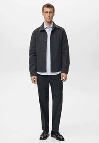 Giacca grigio scuro con design con colletto, chiusura a bottoni e texture morbida, indossata sopra una camicia a righe azzurre e abbinata a pantaloni neri.