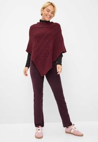 Burgunderfarbener gestrickter Poncho mit Zopfmuster, hochgeschnittener Kragen und lockerer Passform. Kombiniert mit eng anliegenden burgunderfarbenen Hosen und rosa Sneakern.