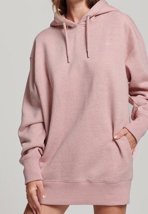 Mujer con sudadera de capucha color rosa claro de gran tamaño, con bolsillo frontal y cordones, posando contra un fondo liso.