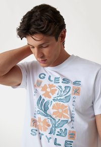 Witte katoenen t-shirt met een kleurrafloraal graphic met blauwe en oranje accenten, ronde halslijn en korte mouwen.