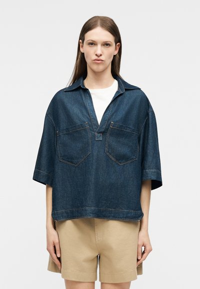 Γυναίκα που φοράει μια άνετη κοντομάνικη μπλούζα από σκούρο τζιν με μπροστινές τσέπες πάνω από λευκό τοπ, συνδυασμένη με μπεζ σορτς μέχρι το γόνατο.