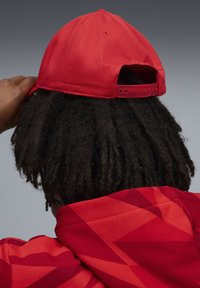 Czerwona czapka baseballowa o gładkiej fakturze z regulowanym paskiem, noszona na odwrót. Częściowo widoczna jest czerwona bluzka z kapturem w wzory pod nią.