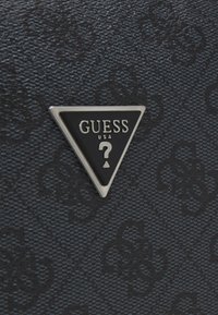 Tessuto nero strutturato con un logo triangolare lucido che presenta "GUESS" e un punto interrogativo, accenti argento e uno sfondo a leggero motivo.