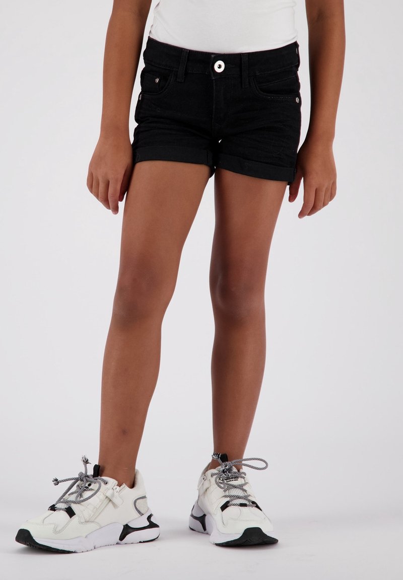 Shorts de mezclilla negra con dobladillos enrollados, que cuentan con bolsillos frontales y un botón brillante. Combinados con zapatillas blancas gruesas con acentos negros.