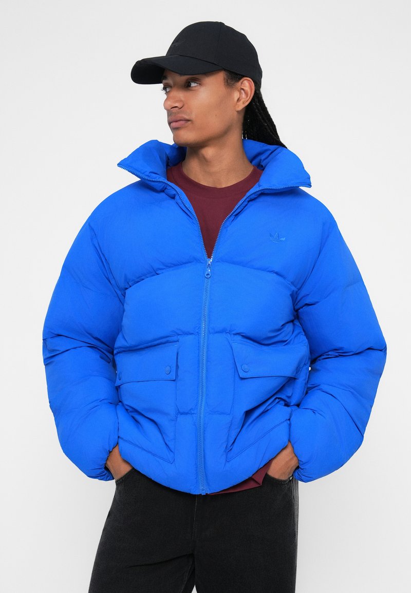 Giacca puffer blu royal con colletto alto, zip frontale e due tasche anteriori. Presenta un tessuto liscio e strutturato. Cappello nero indossato con essa.