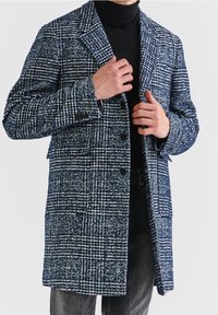 Marineblauwe en witte geruite wollen jas met revers, knoopsluiting en twee zakken aan de voorkant, gedragen over een zwarte coltrui en spijkerbroek.