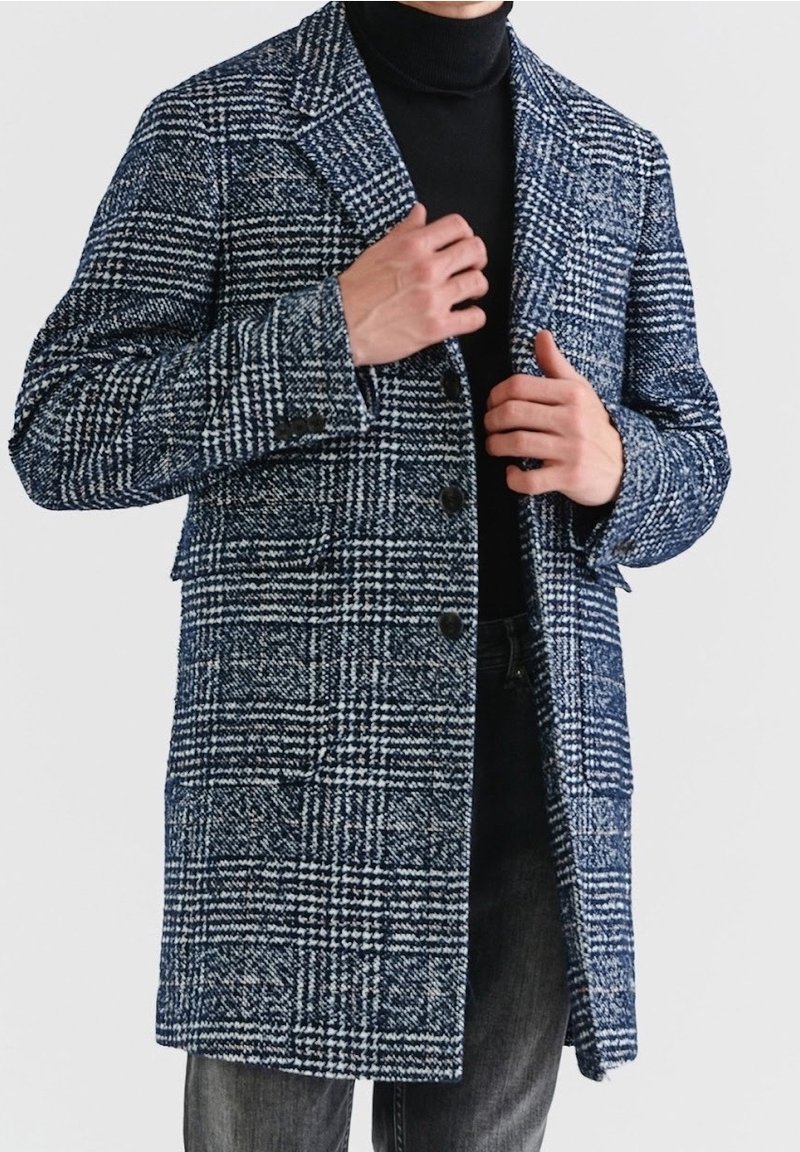 Marineblauwe en witte geruite wollen jas met revers, knoopsluiting en twee zakken aan de voorkant, gedragen over een zwarte coltrui en spijkerbroek.